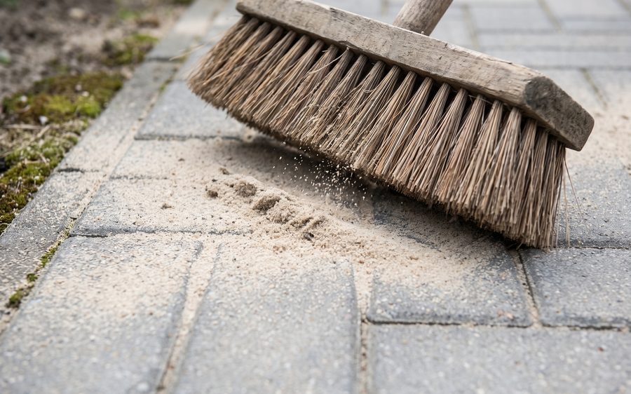 A yard broom sweeping pale sand into the joints of cleaned block paving